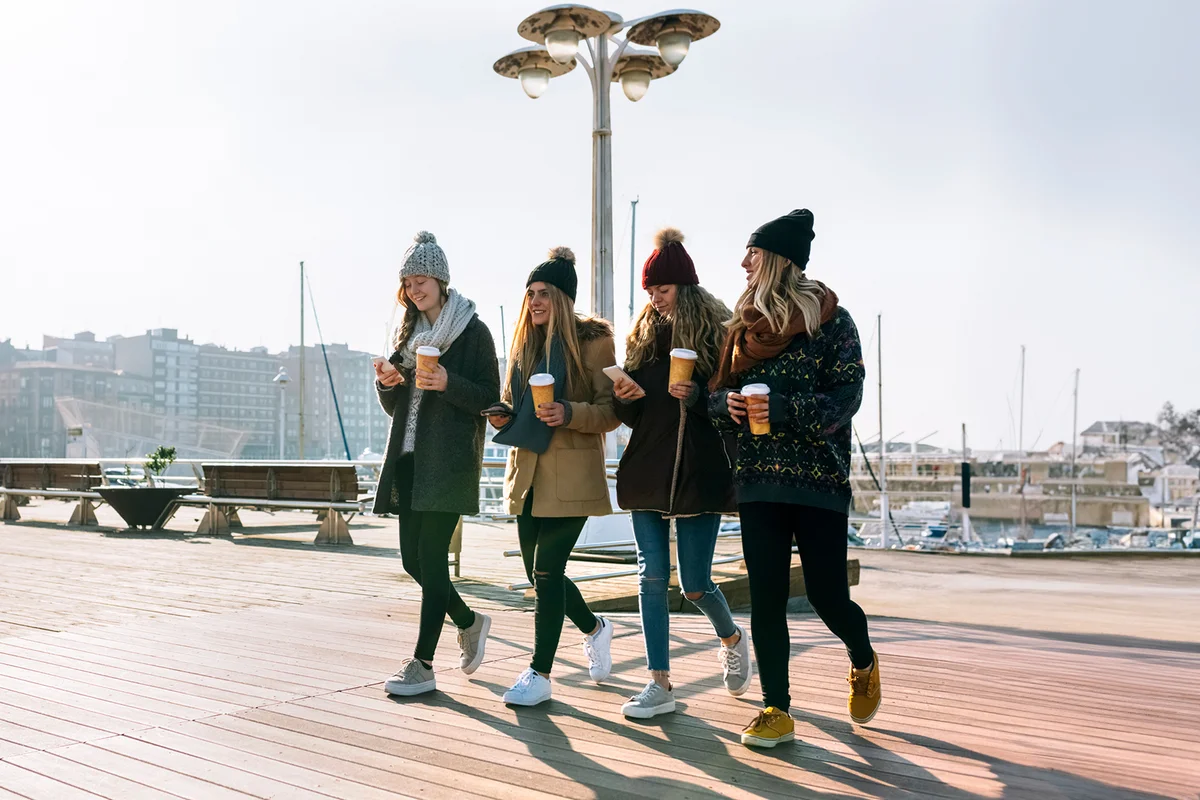 Four friends with coffee to go and cell phones walking on promenade in winter