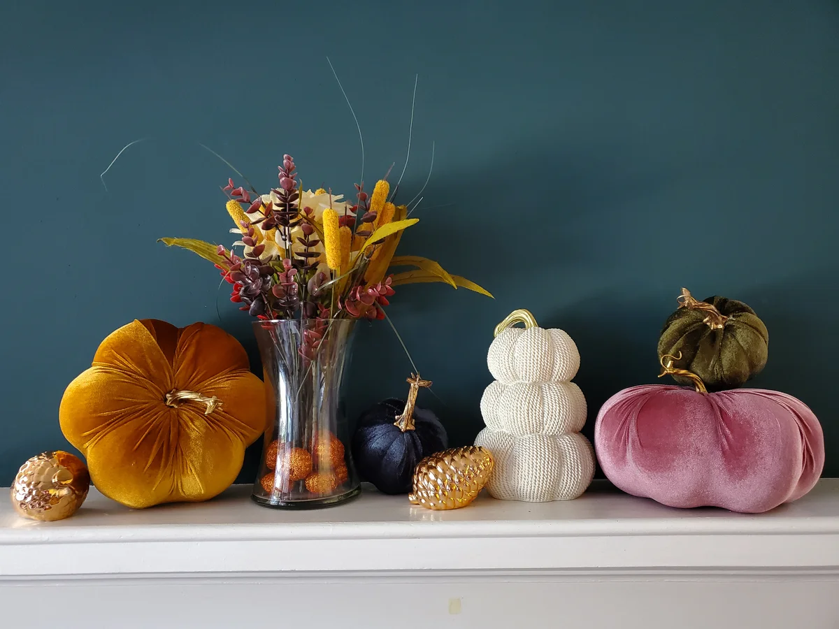 A decorated fall mantel with two large velvet pumpkins and a full floral arrangement. Smaller pumpkins and ceramic figures of a pinecone and acorn complete the look.