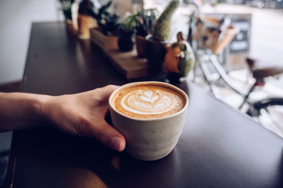 Cropped shot of someone hand with a cup of hot latte coffee on the table nearly window