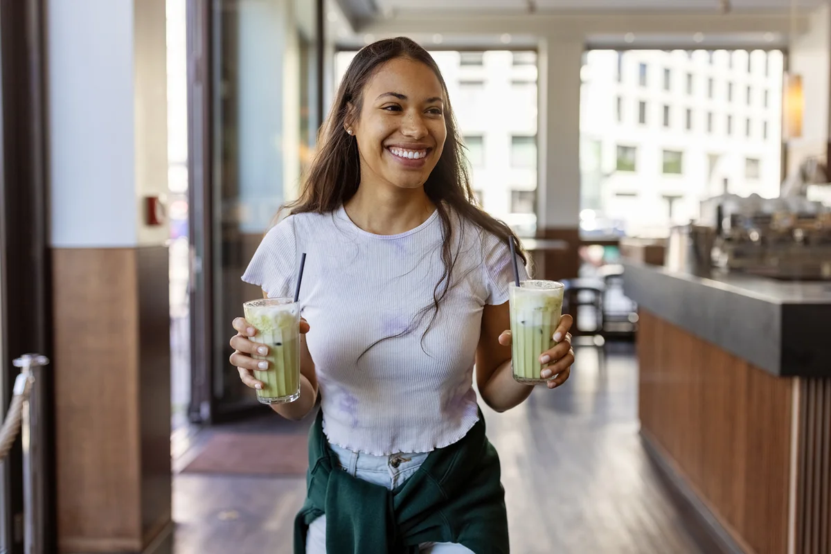 Happy young woman with two cold matcha latte glasses inside city cafe