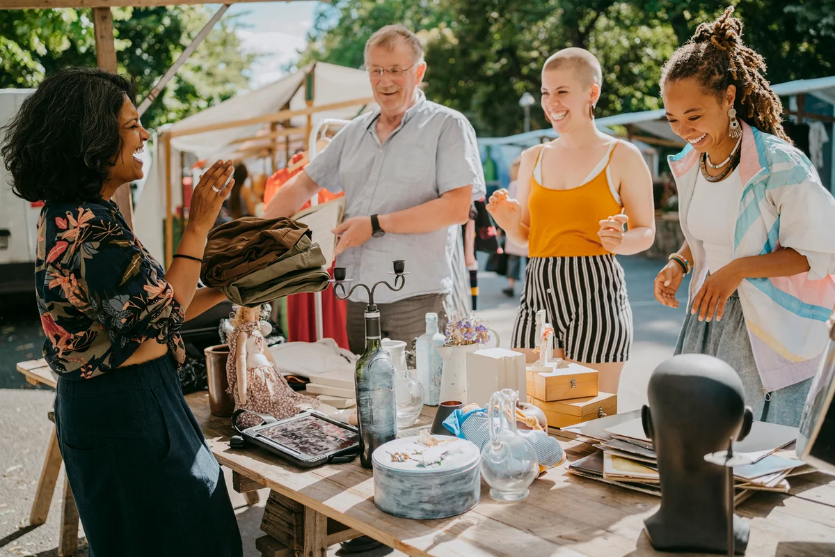 happy female owner holding dress with customers shopping near stall at flea market