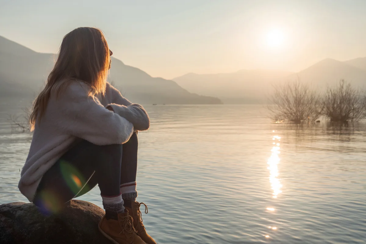 Woman contemplates lake at sunset from boulder