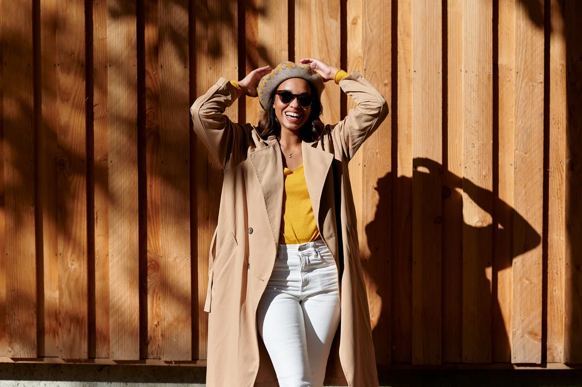Woman in trench coat raising arms in front of wooden wall