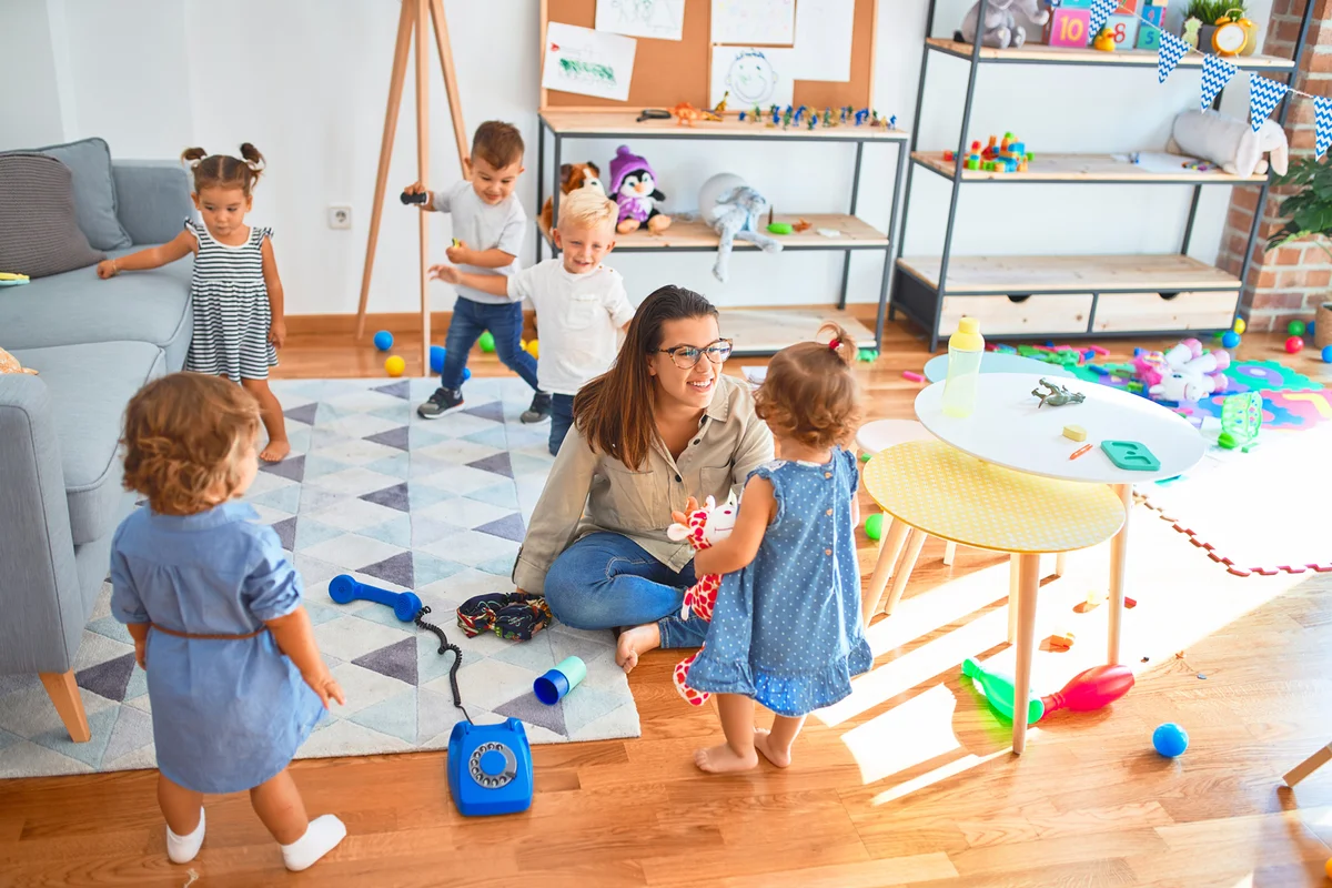 teacher and group of toddlers playing around lots of toys at kindergarten