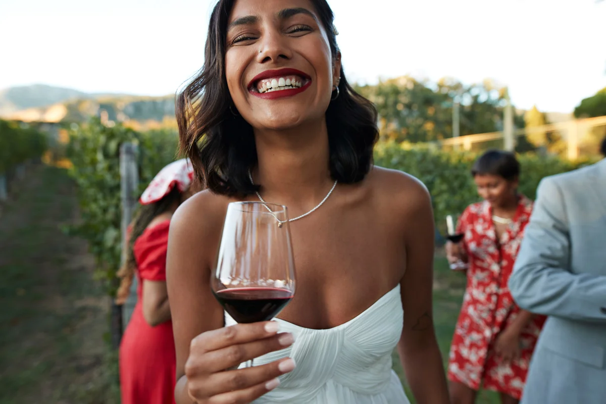 Portrait of laughing young bride in off shoulder wedding dress holding wineglass at vineyard