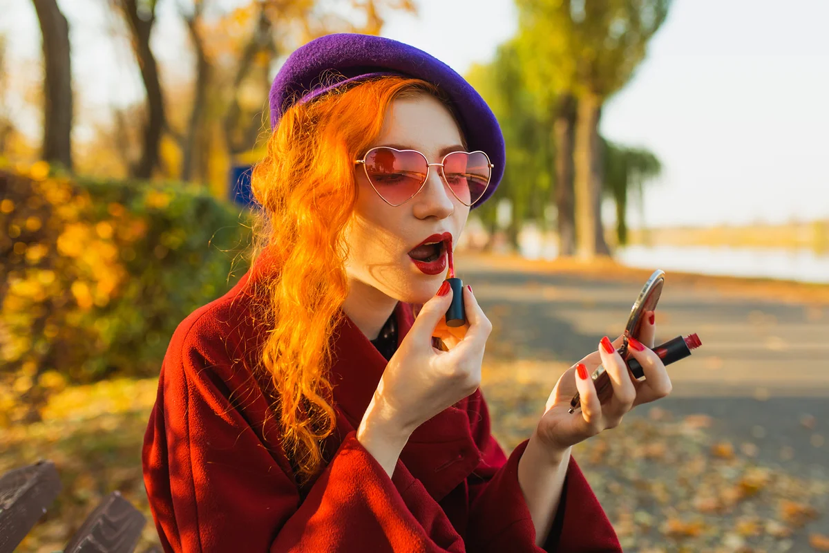 Stylish redhead woman in red coat paints lips with gloss
