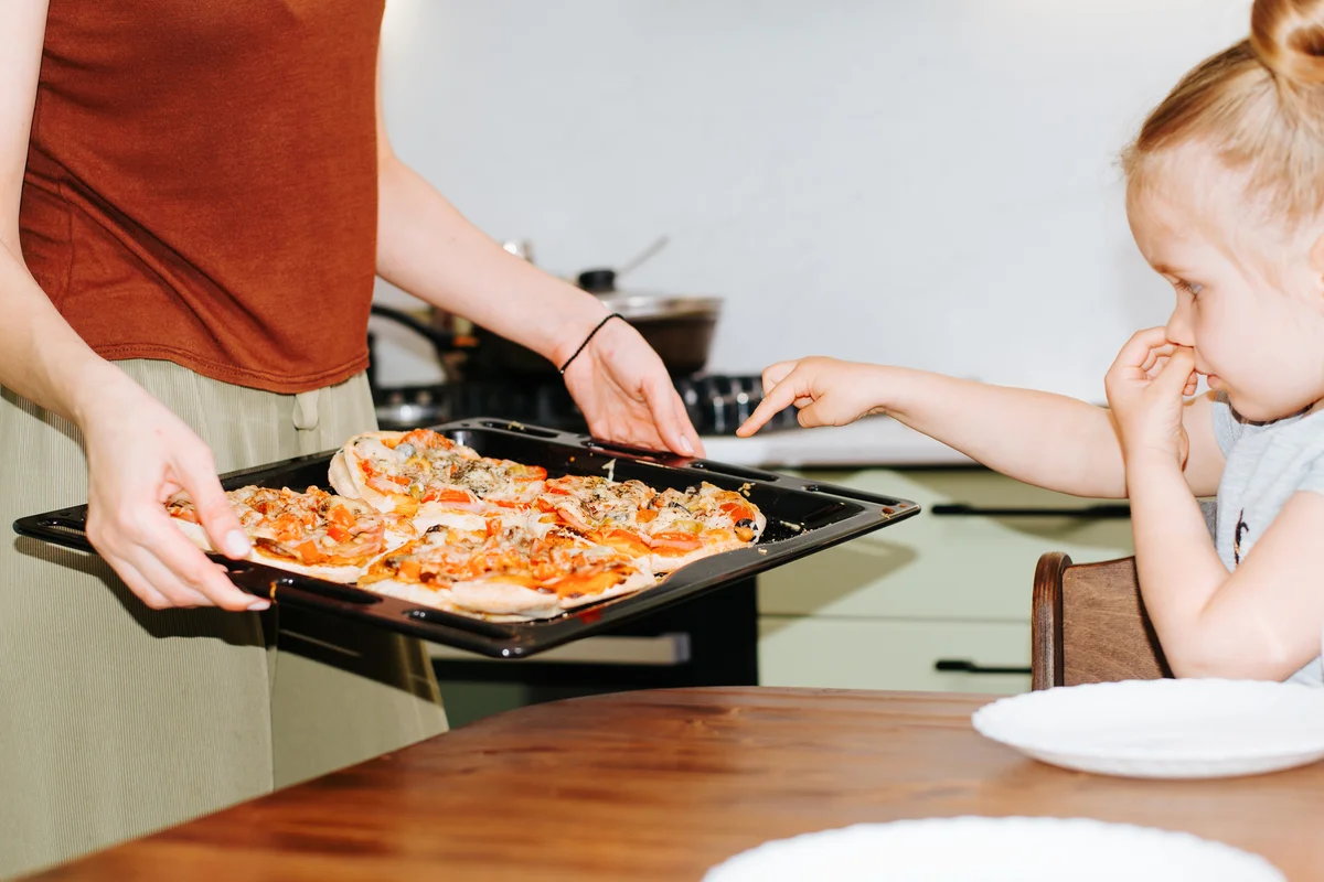 daughter choosing mini pizza, close-up mom showing cooked food to child in kitchen.