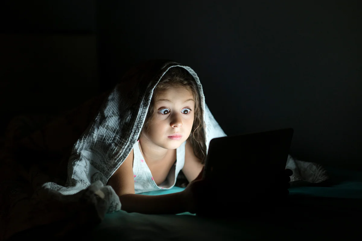 Little girl reading funny ghost stories tablet in her bed at night