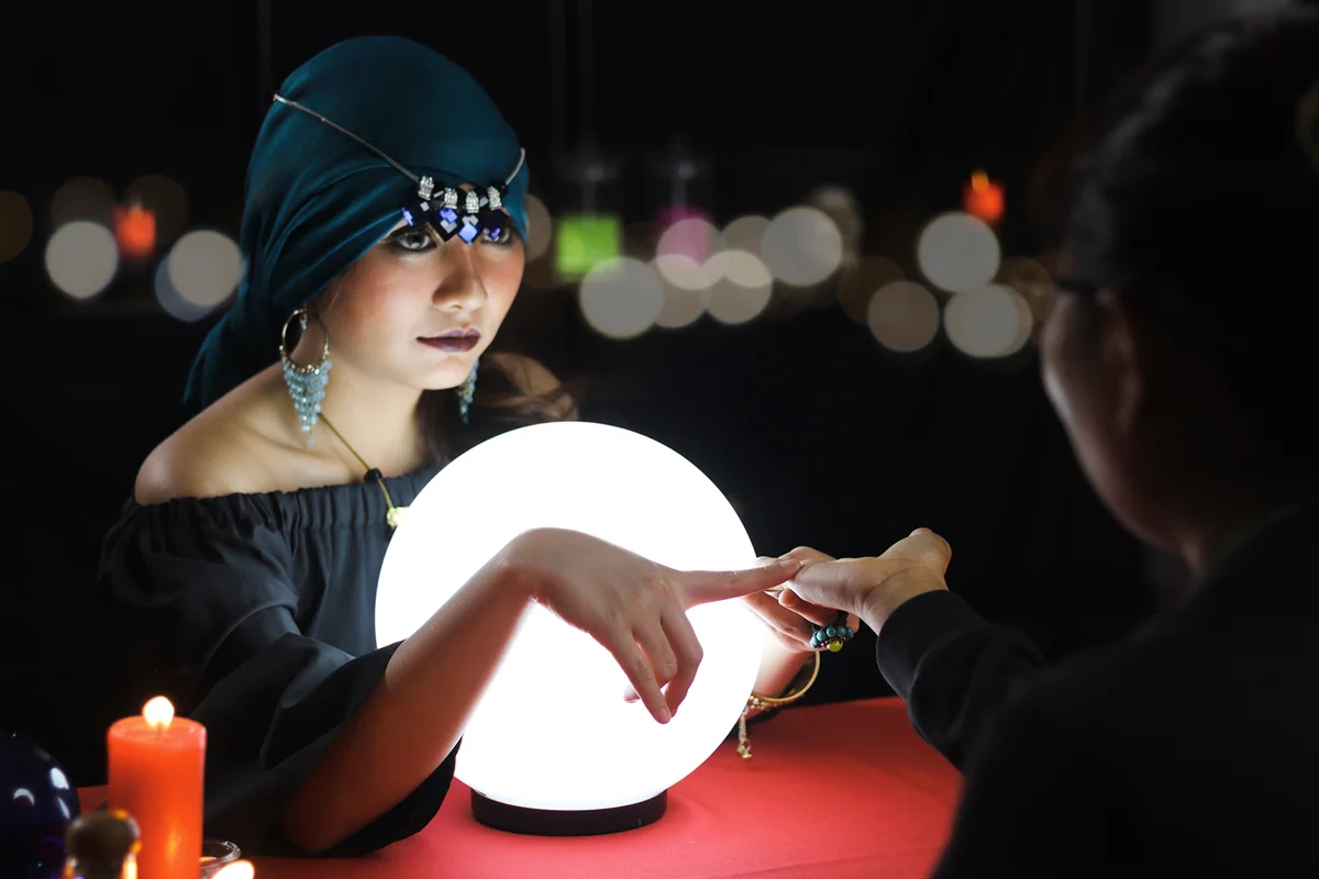 Mysterious fortune teller in black dress with magical crystal ball pointing to the customer palm