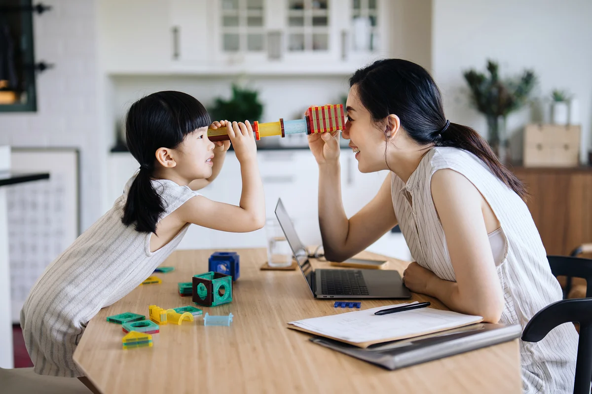 mother working from home on laptop while little daughter playing with her on the desk