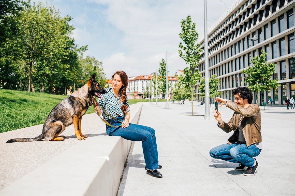 Man photographing woman sitting by dog through mobile phone in city