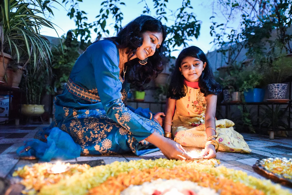 Mother and daughter lighting lamps around rangoli made using petals