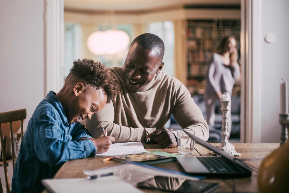 Son writing while studying by father over table at home