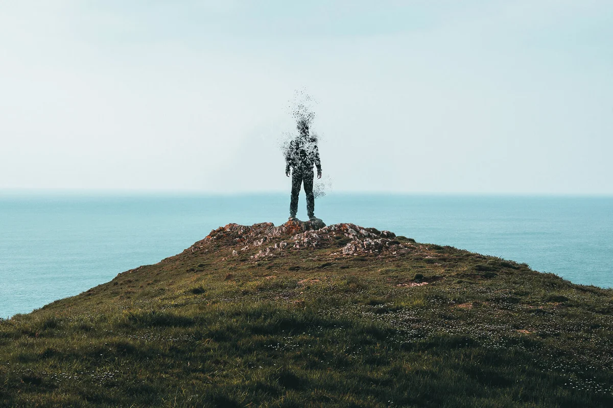 A lone hooded figure standing on top of a hill and disintegrating while looking out across the ocean