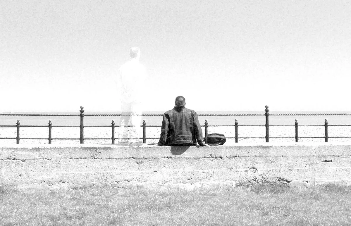 Rear View Of Man Sitting By Ghost Standing On Retaining Wall Against Clear Sky