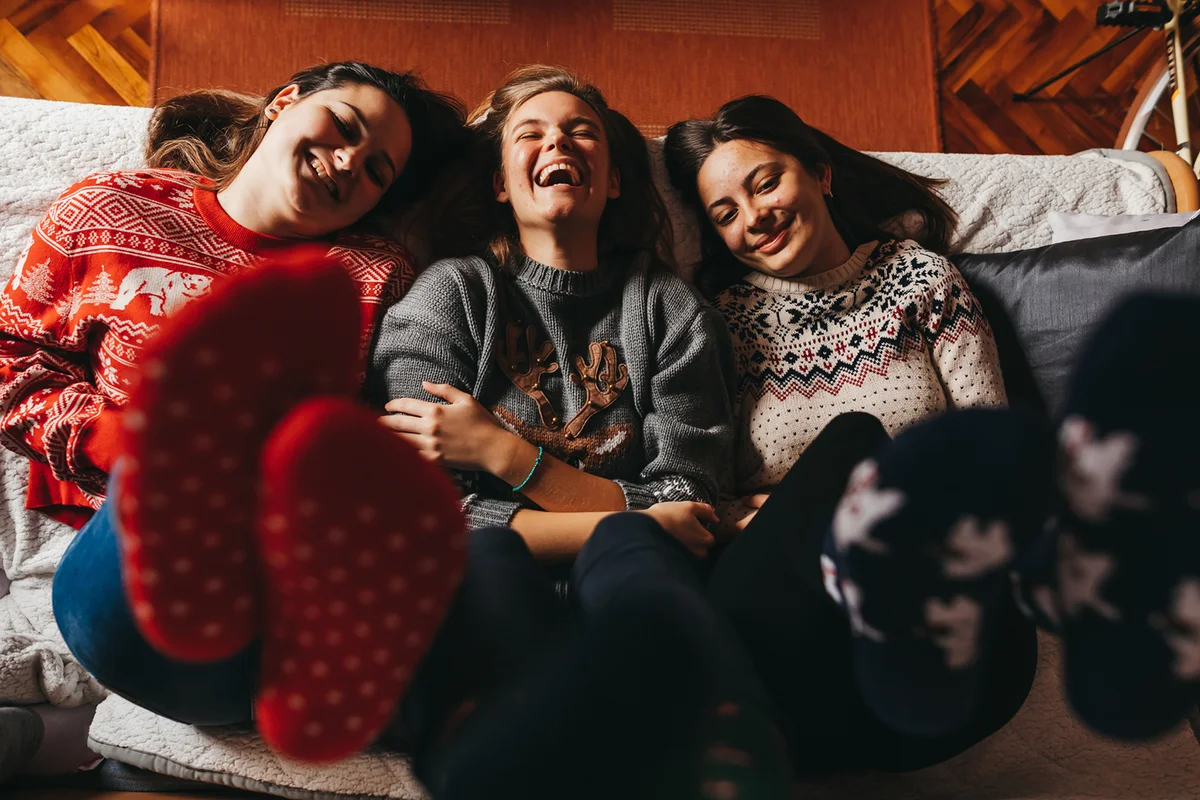 Three young cheerful female friends in Christmas sweaters and socks lying on sofa with feet up at home