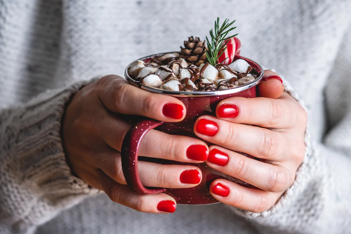 A woman with red nails holding a cup of hot chocolate decorated with marshmallows, red candy and a Christmas tree branch
