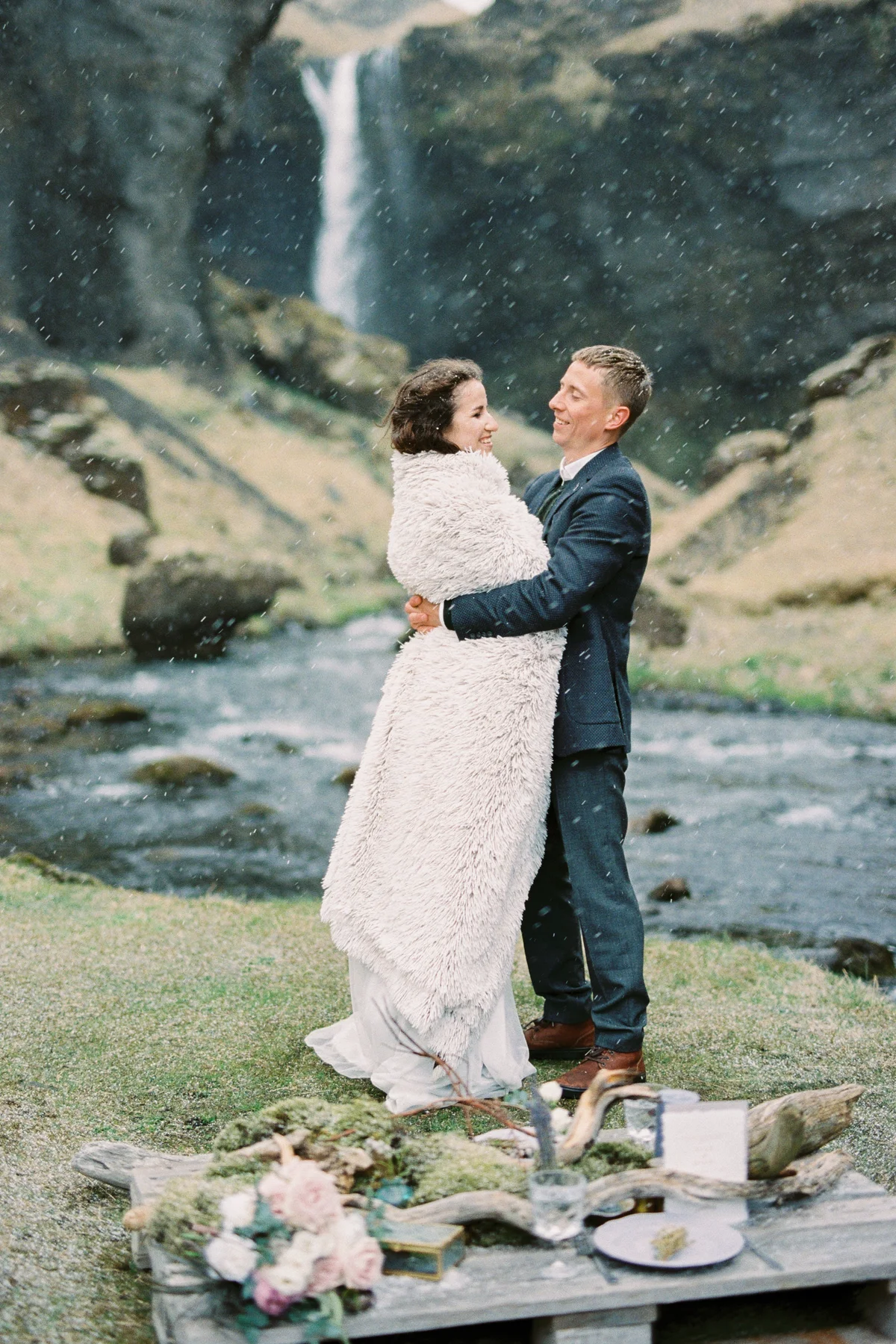 Groom hugs bride in a blanket on the river bank