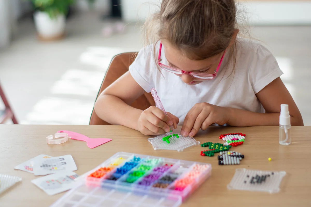 A girl sitting at a table makes figures out of colorful beads. Aqua mosaic game.