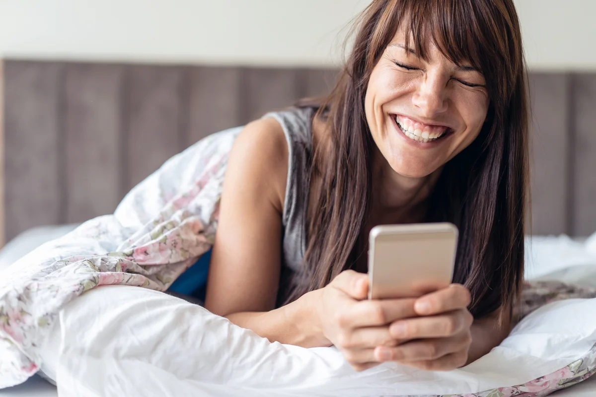 Woman laying in the bed holding smart phone and laughing