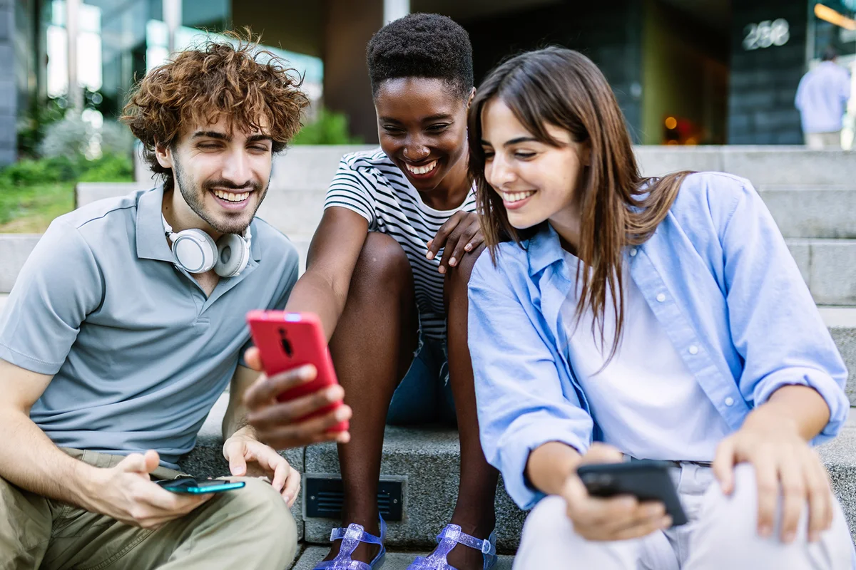 Multiracial young adult people having fun watching funny social media content on smartphone app while relaxing together outdoors