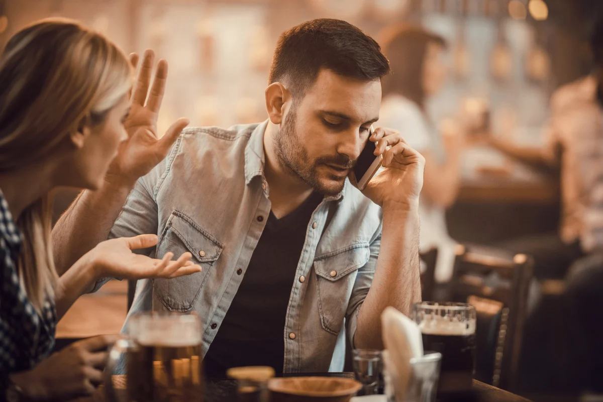 Young man talking on cell phone while gesturing to his date to stop talking
