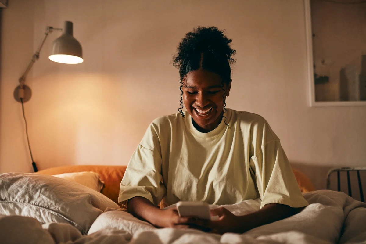 Happy teenager girl using mobile phone sitting on bed at home