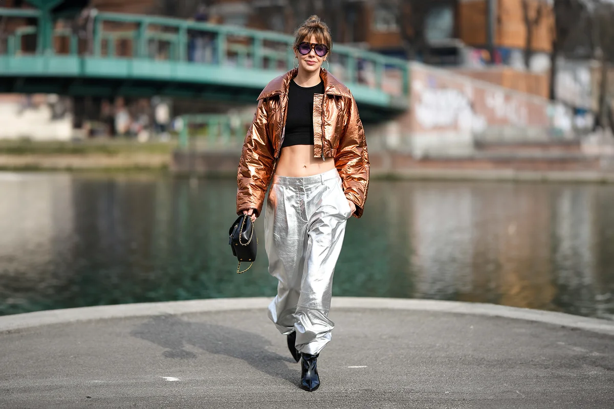 Woman wearing a brown shiny metallic shiny leather cropped jacket, silver shiny metallic leather puffy pants, a black shiny leather handbag