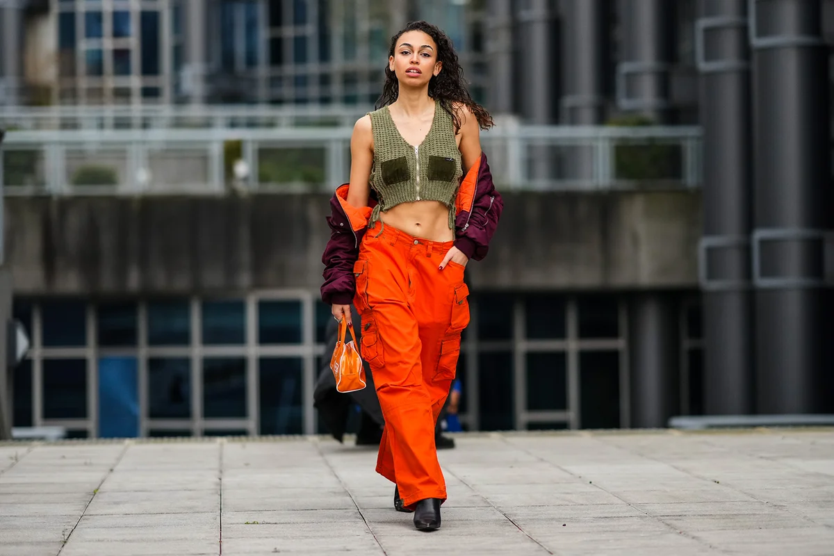 Maria Decremps wears earrings, a khaki wool braided V-neck / cropped / zipper tank-top, a purple cropped bomber coat, high waist orange cargo pants, an orange shiny leather with a white "Ciao-Bella" inscription print pattern handbag