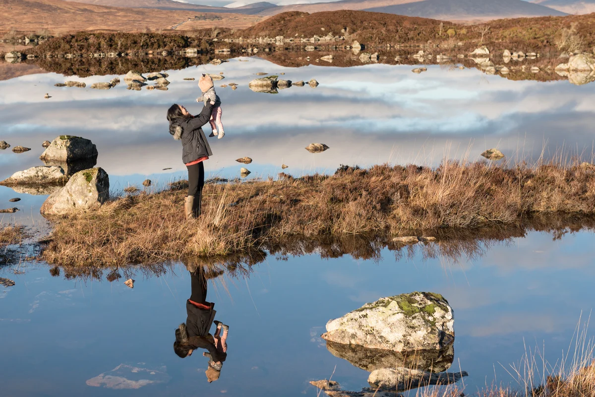 Woman stands on marsh and holds her baby up in the Loch Etive