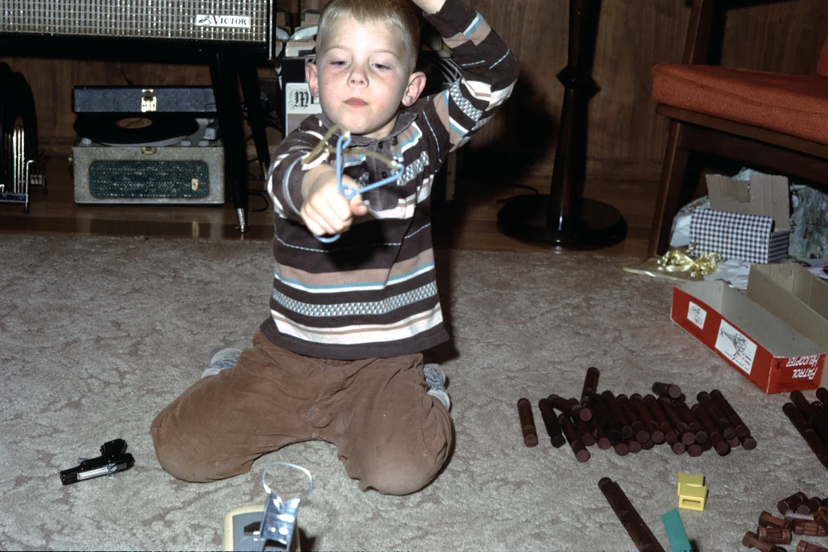 Young boy playing with a slingshot, Lincoln Logs, and a toy gun in a suburban living room, United States, 1961