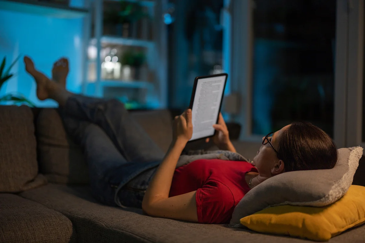 Woman lying on sofa in living room at home, and reading a book on digital tablet in peace