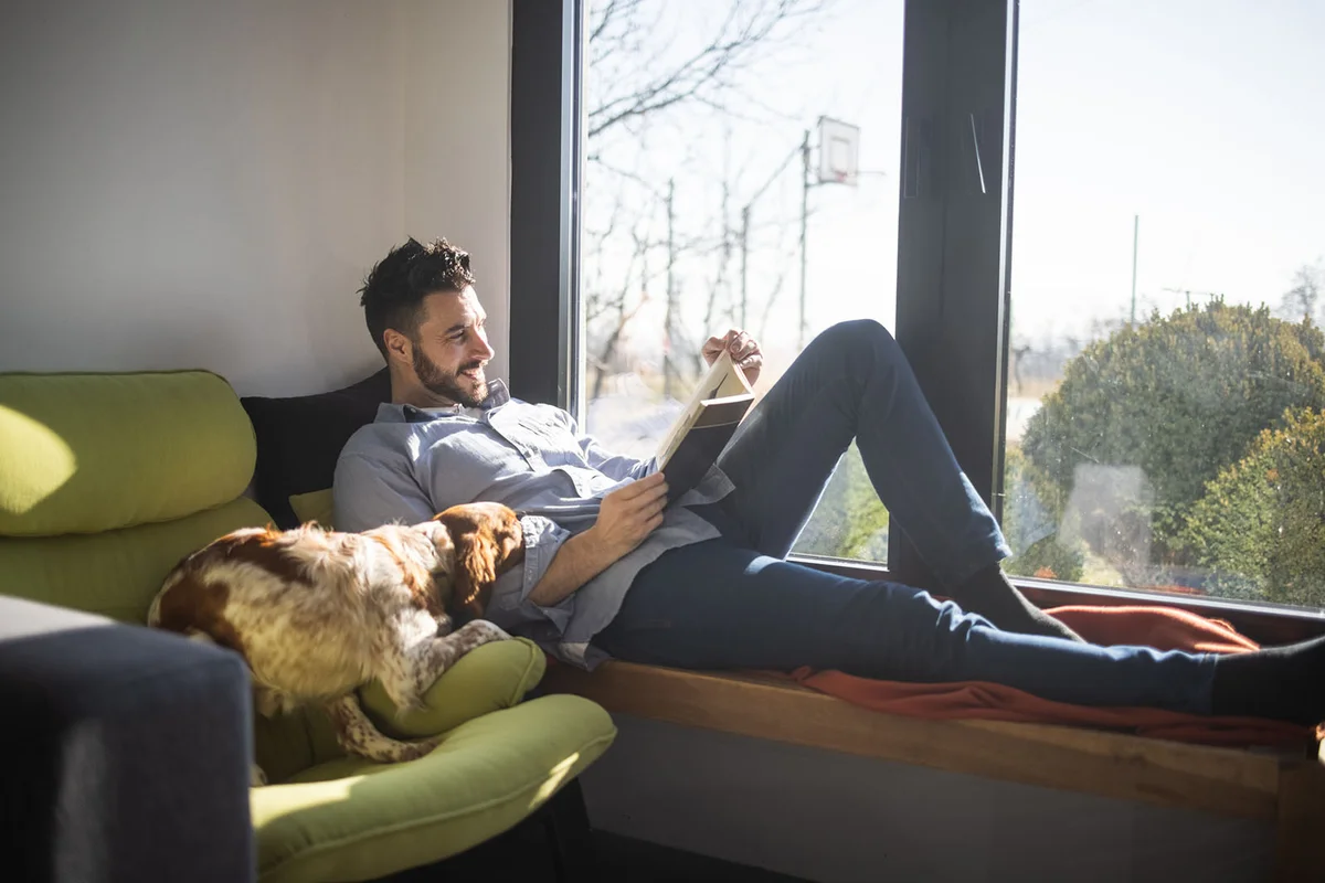 man lying on bench next to window and reading book