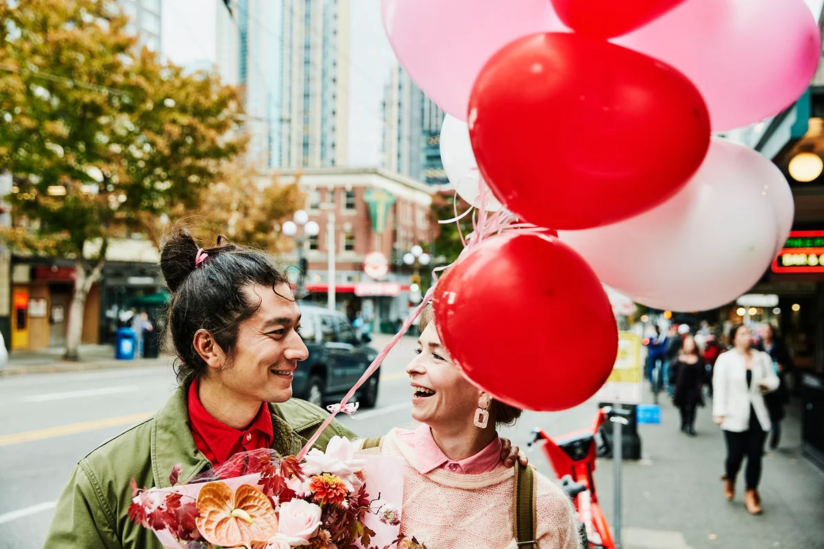 Smiling couple walking down city sidewalk with flowers and balloons during Valentines date