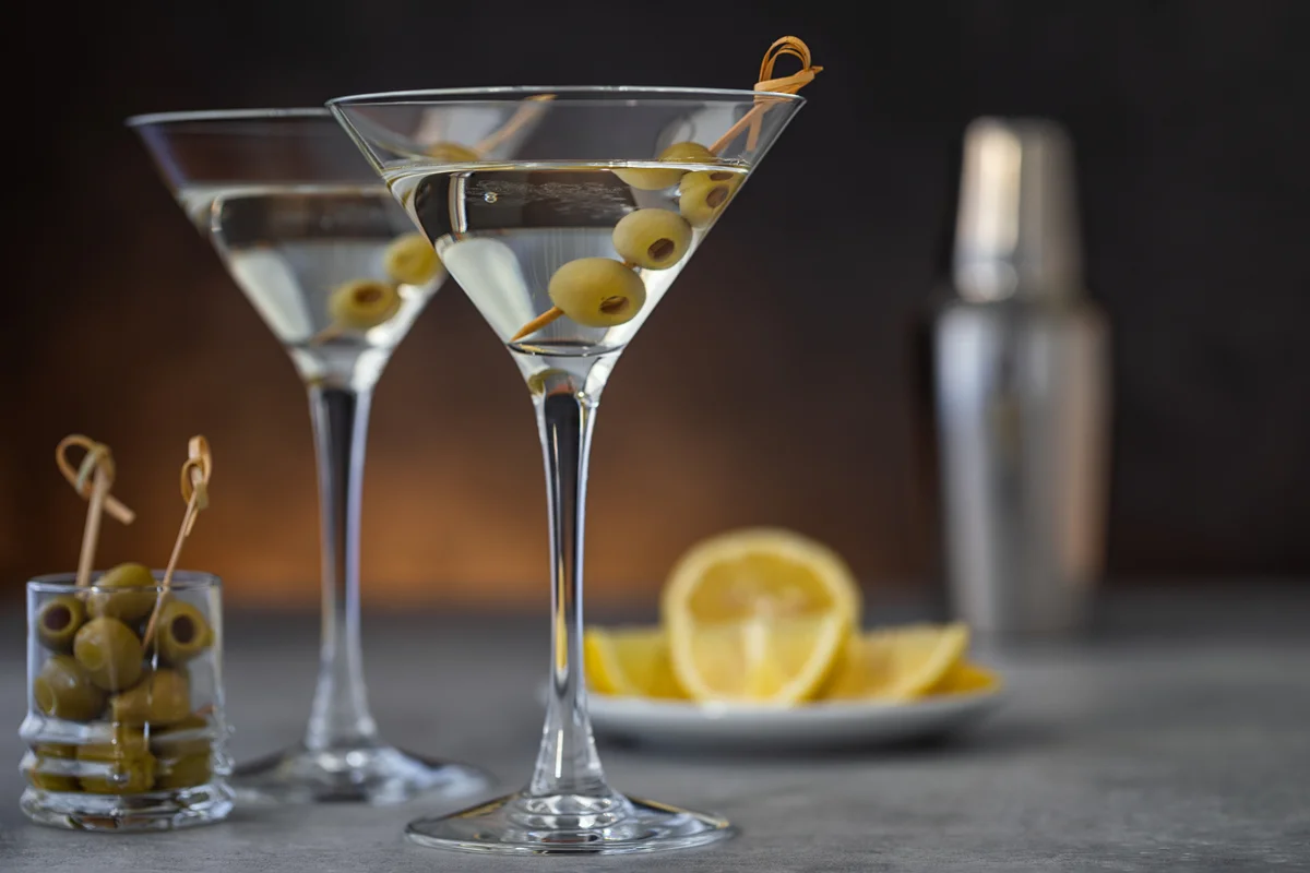 Two martinis with olives and bartending paraphernalia around them.