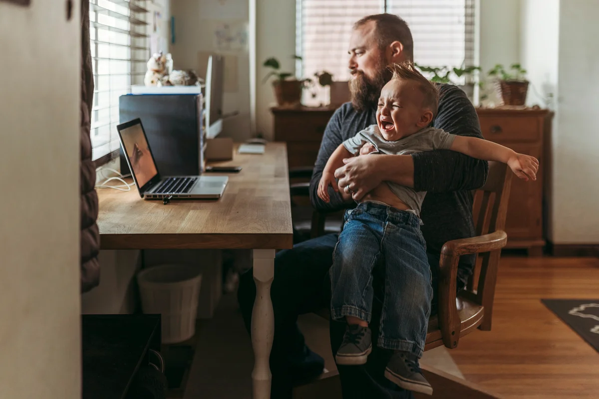 Dad working from home with one year old boy crying in his lap