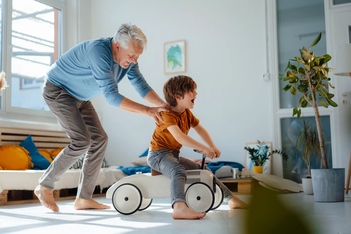 Playful senior man pushing grandson sitting on toy car in living room at home