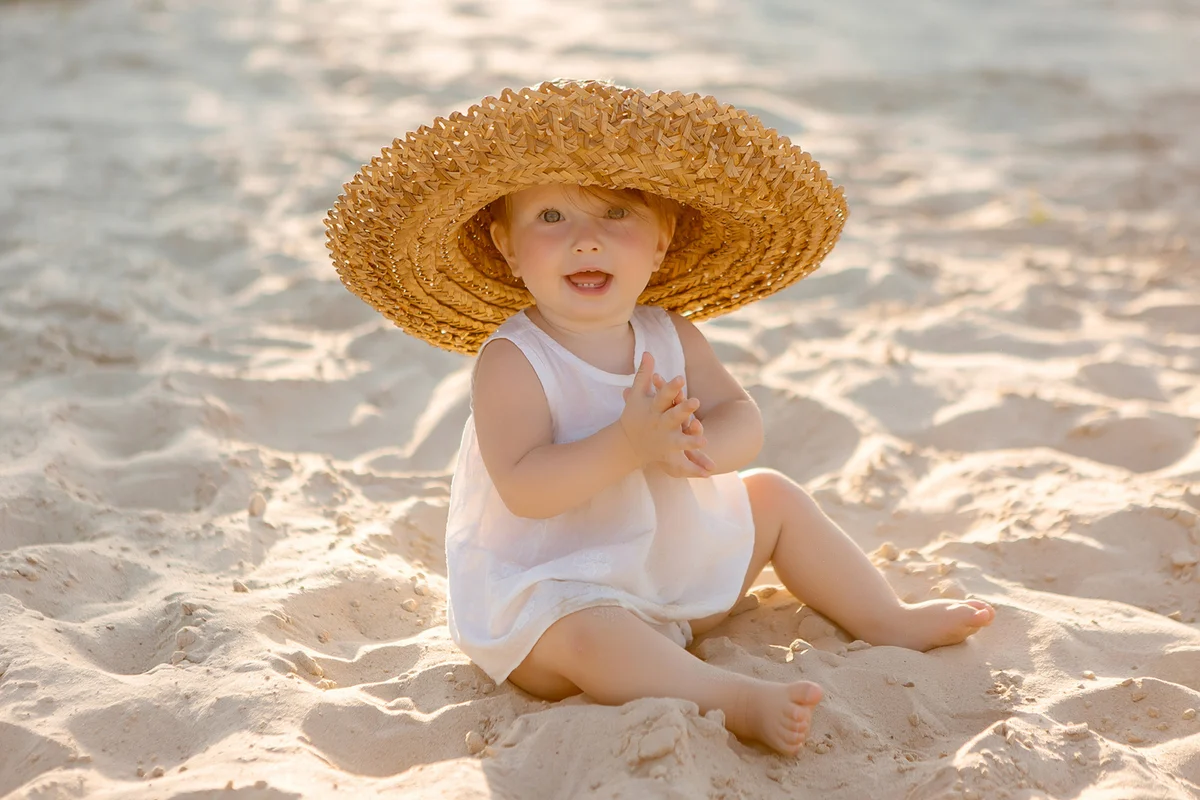 Baby girl in white clothes and a straw hat sits on the white sand on the beach in summer