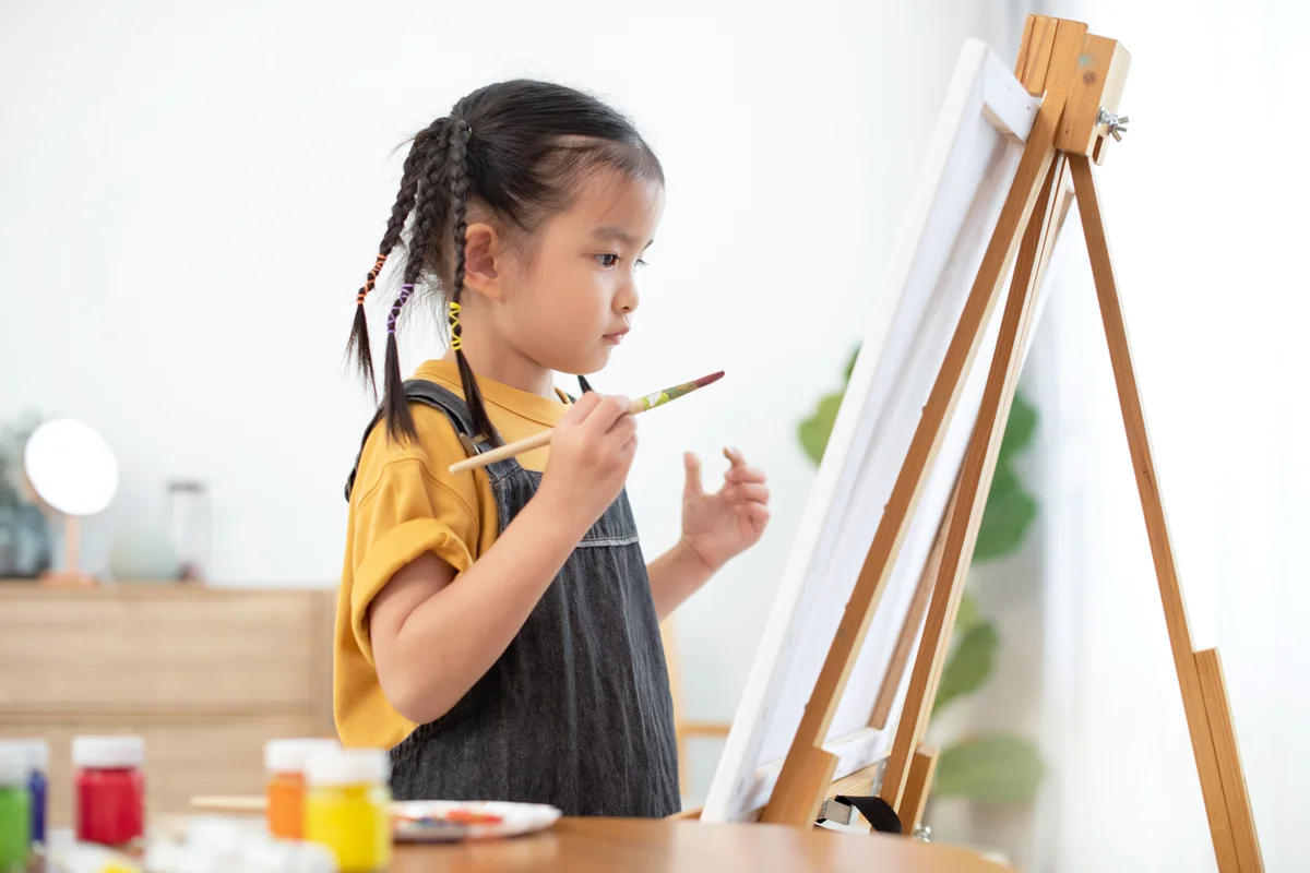 Little girl painting in canvas standing on easel at home