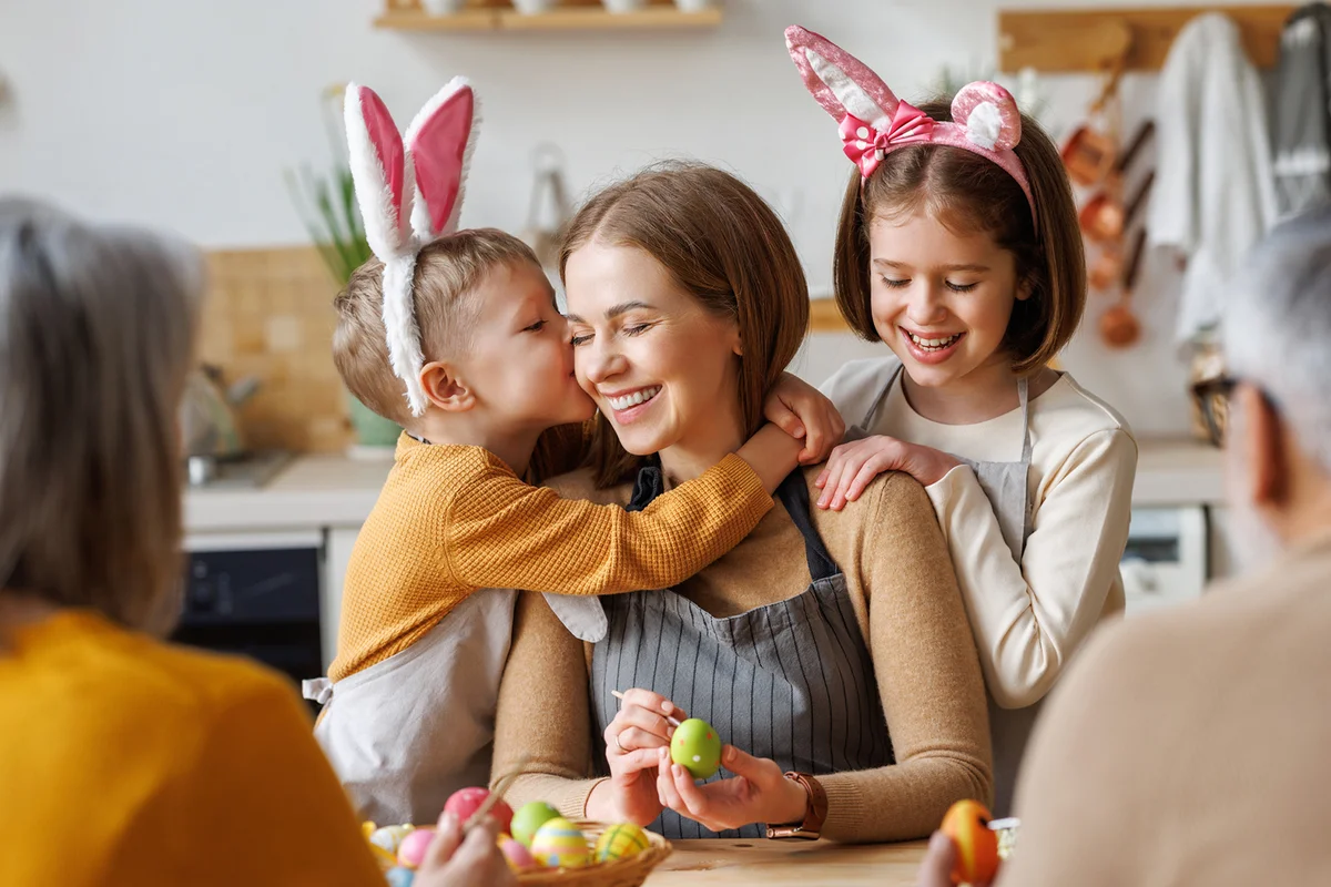 Cute little children embracing and kissing young happy mother while painting Easter eggs with family