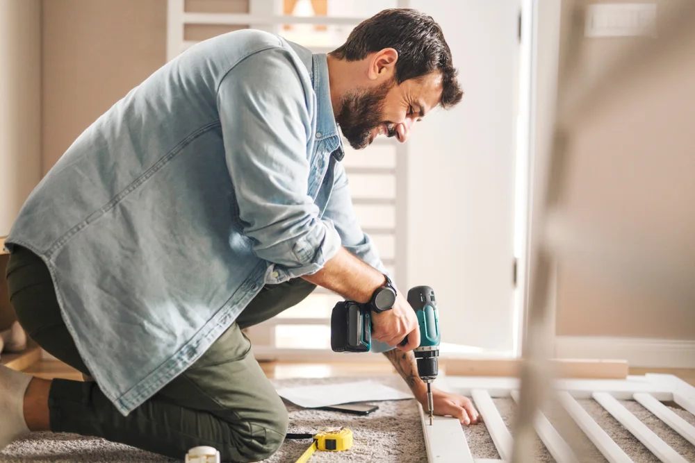 A smiling man holding screwdriver machine and screwing a slats of a baby crib