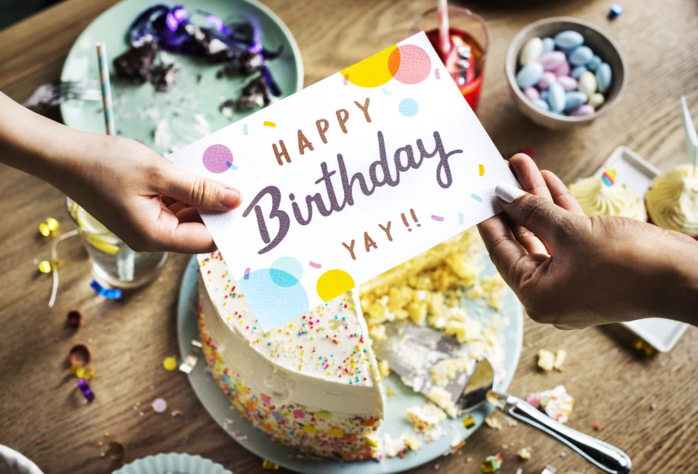 Someone's hand receiving a birthday card over a table with birthday cake and plates