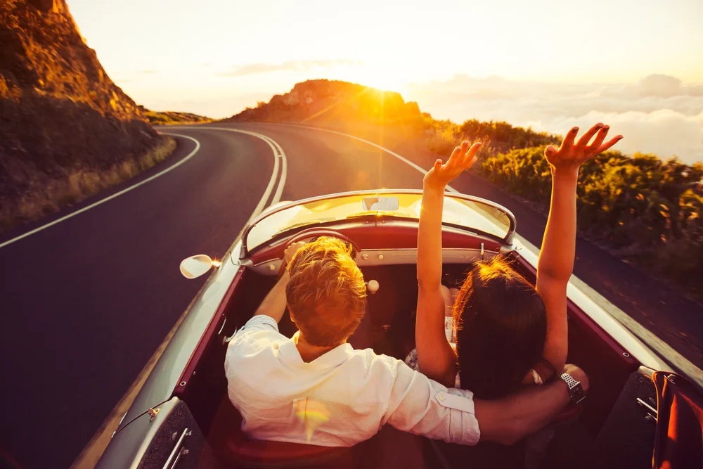 Couple driving a vintage convertible car with beautiful mountain and cloud views at sunset