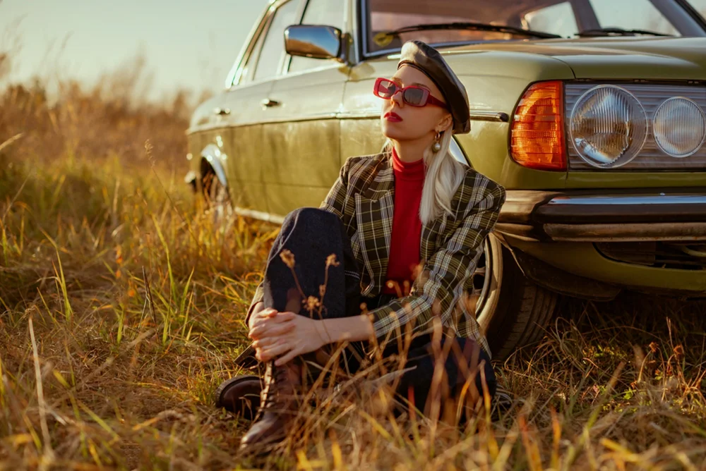 Stylish woman sitting against her vintage car in a field