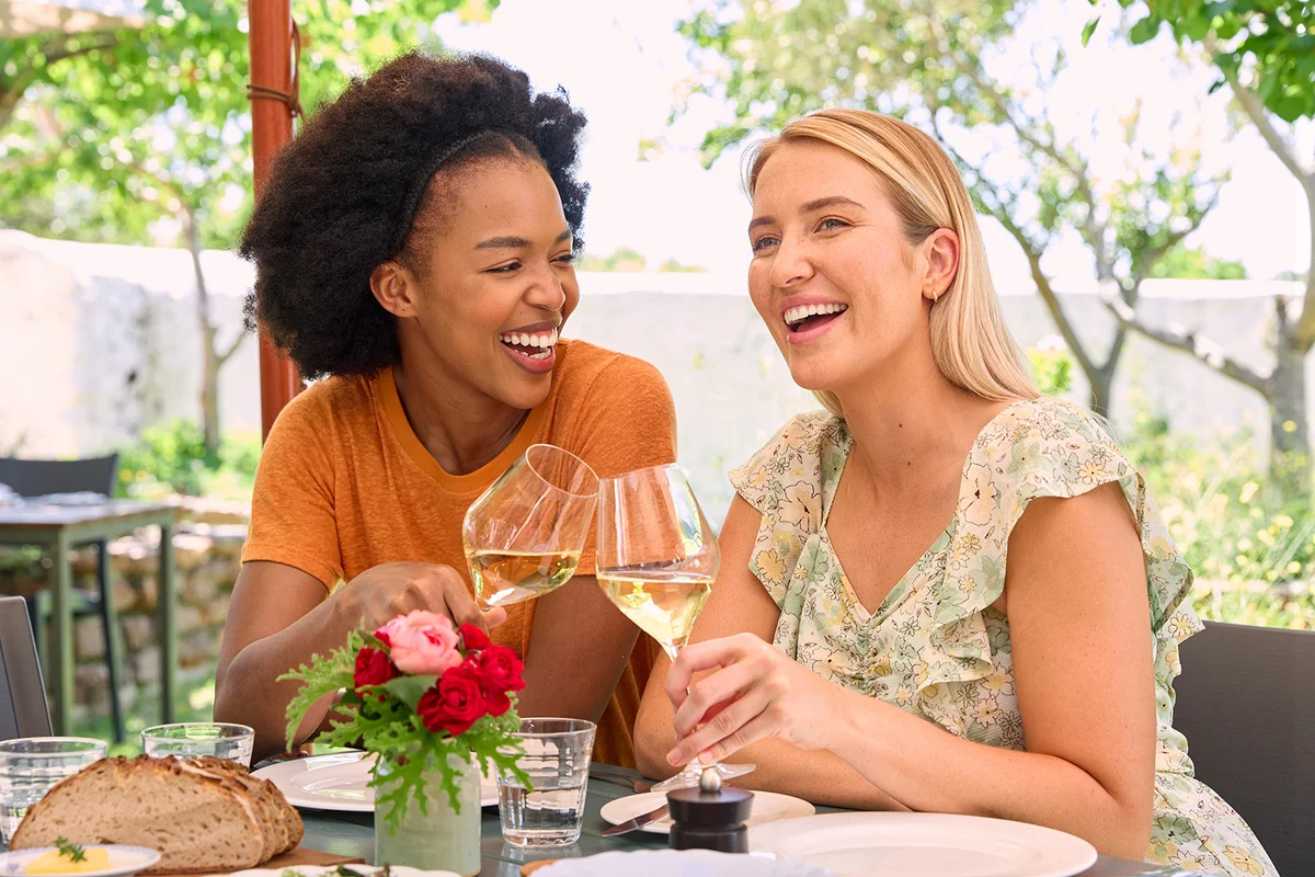 Two Female Friends Enjoying Outdoor Meal And Wine On Visit To Vineyard