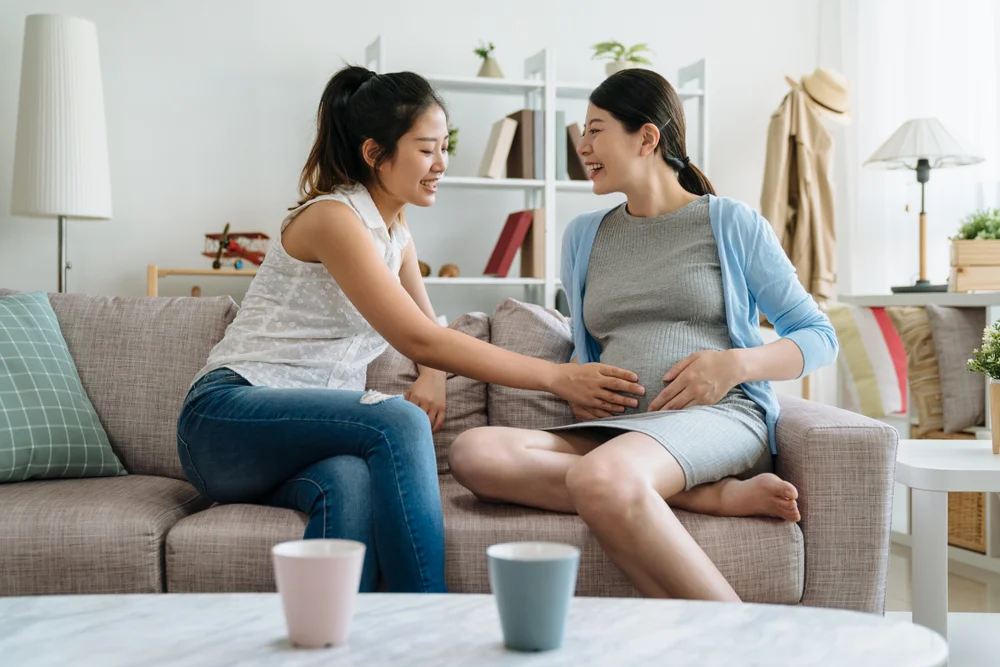 Woman touching her pregnant friend's belly at home