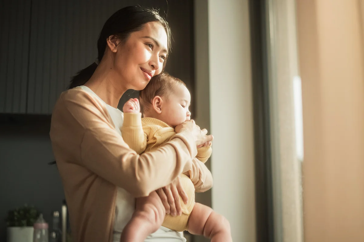 Mother Holding her Baby While Standing Next to a Window at Home