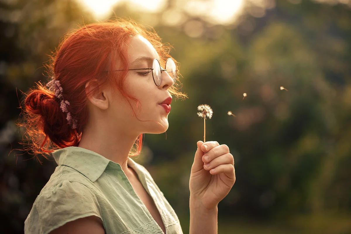 redhair curly girl wearing glasses blowing dandelion