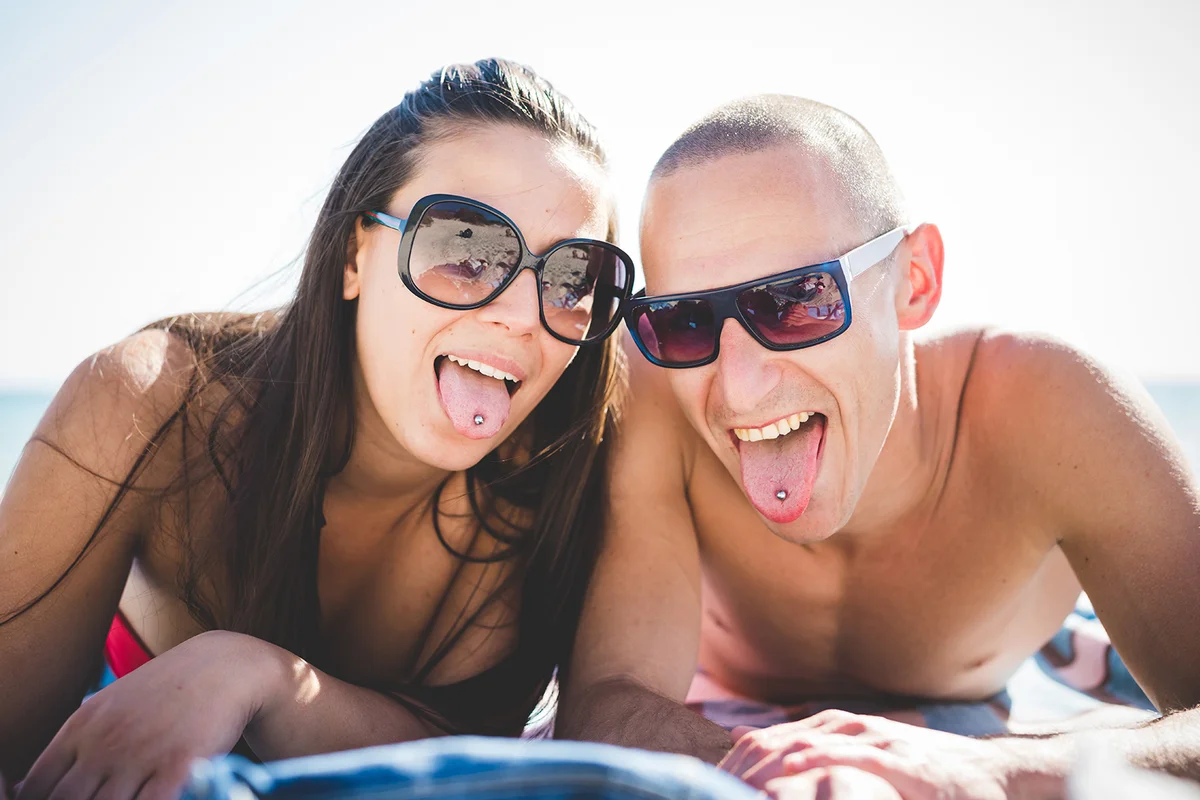 couple lying on beach toowl in summertime using smartphone showing tongue piercing