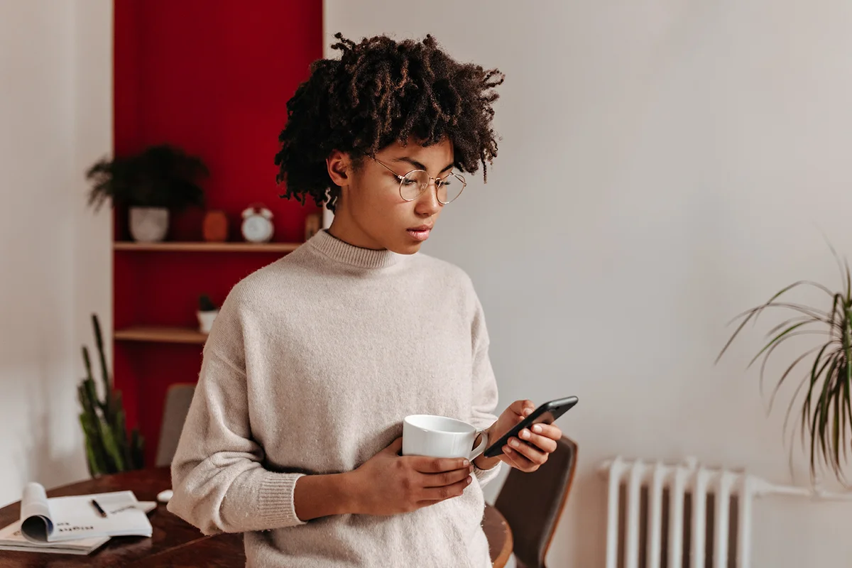 Girl in beige sweater is looking intently at phone and posing in office with white cup